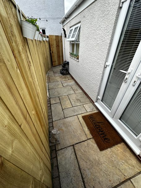 Narrow stone garden path beside a wooden fence leading to a door with a welcome mat.