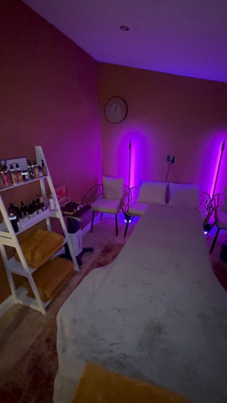 A serene treatment room with purple lighting, a massage table, and wooden shelves.