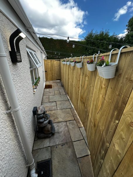 Narrow garden path with wooden fence, planters, and a decorative statue.